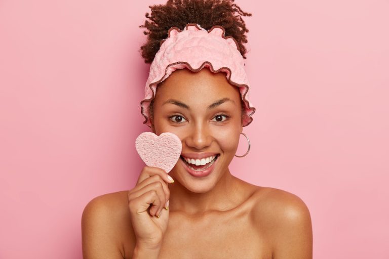 Headshot Of Beautiful Dark Skinned Woman Has Healthy Skin, Wipes Face With Cosmetic Sponge, Smiles Positively, Shows White Teeth, Wears Shower Headband, Takes Shower After Work, Isolated On Pink