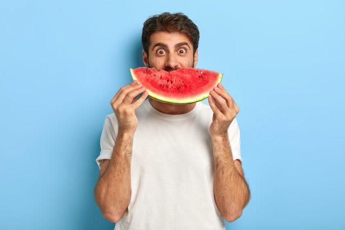 Funny Man Holds Half Of Red Watermelon Near Face, Has Glad Surprised Look, Dressed Casually, Has Healthy Summer Nutrition. European Guy Poses With Tasty Tropical Fruit Against Blue Background
